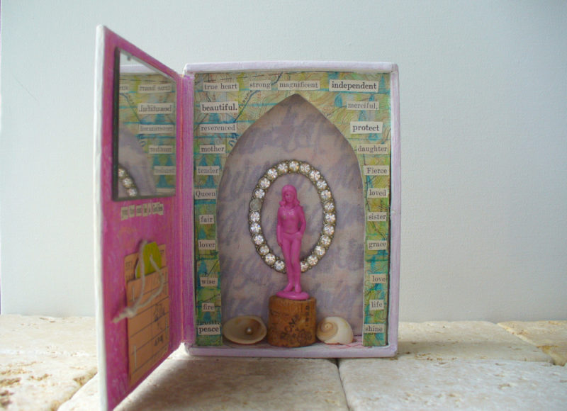 goddess shrine made in a cigar box with pink plastic toy, rhinestone pin, mirror, shells and words