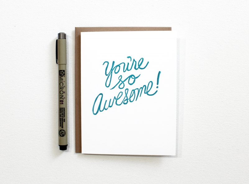 block printed you're so awesome note card hand printed in script in turquoise ink on a white card with a kraft envelope shown with a pen for size